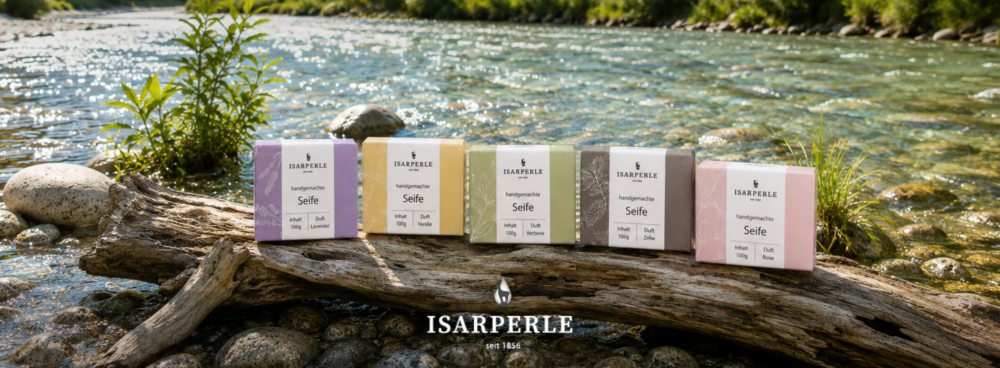 Isarperle Markenshop-Banner – Fünf handgemachte Seifen à 100g auf einem Treibholzstamm an einem klaren Gebirgsfluss: Duft Lavendel, Vanille, Verbene, Zirbe und Rose. Isarperle seit 1856.
