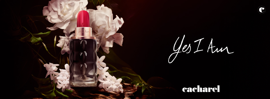 Werbung für das Parfum 'Yes I Am' von Cacharel. Im linken Bildbereich steht ein Lippenstift-förmiger Parfumflakon mit roter Kappe und dunklem Glasdesign, umgeben von weißen Blüten. Rechts im Bild steht in handschriftlicher Schrift 'Yes I Am' und darunter das Cacharel-Logo. Der Hintergrund ist dunkel, wodurch der Flakon und die Blumen dramatisch hervorgehoben werden.