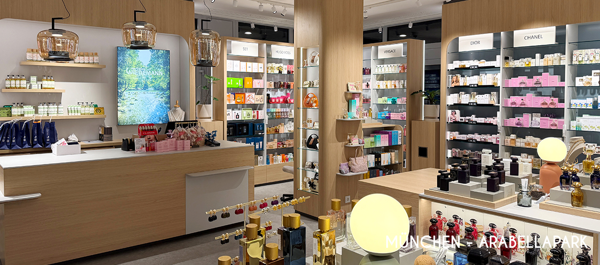 Parfümerie Filiale München-Arabellapark – Innenansicht mit Kassenbereich, Bildschirm-Werbedisplay, Schmuck- und Taschen-Vitrinen sowie Regalen für Dior und Chanel Parfums.