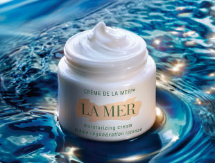 Werbebild für ‚The Moisturizing Fresh Cream‘ von La Mer. Ein geöffneter Tiegel mit Creme steht auf einer nassen, glitzernden Wasseroberfläche vor einem blauen Hintergrund. Links daneben steht der Text: ‚Die neue The Moisturizing Fresh Cream – Verjüngung für eine erfrischte, ausgeglichene Haut‘.