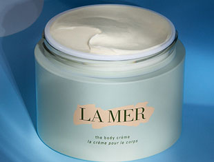 Geöffneter Tiegel der ‚La Mer The Body Crème‘ vor blauem Hintergrund. Die cremige Textur ist sichtbar. Auf dem hellgrünen Tiegel steht: ‚La Mer – the body crème – la crème pour le corps‘.