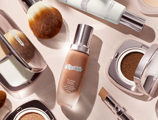 Verschiedene Make-up-Produkte von La Mer auf hellem Untergrund. In der Mitte eine Foundationflasche mit silbernem Pumpspender, umgeben von Puderdosen, Concealer, Pinsel und weiteren Kosmetikprodukten in eleganter Verpackung.