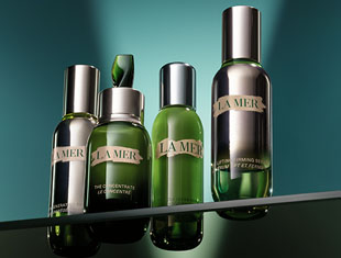 Vier Pflegeprodukte von La Mer in grünen Flaschen mit silbernem Deckel auf einer glänzenden Oberfläche. Von links nach rechts: Pipettenflasche, Pumpspender, Spray und Lotion. Der Hintergrund ist türkisfarben mit Lichtverlauf.