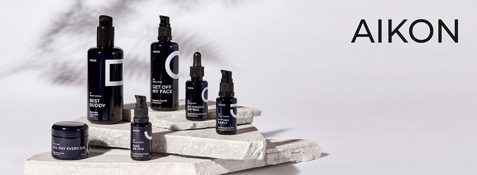 Pflegelinie von AIKON: Mehrere dunkelblaue Flaschen und Tiegel mit weißer Beschriftung stehen auf hellen Steinplatten vor grauem Hintergrund, rechts daneben das Logo „AIKON“.