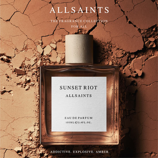 Parfumflakon „Sunset Riot“ von ALLSAINTS liegt auf gebrochener, terrakottafarbener Erde. Darüber steht „ALLSAINTS – The Fragrance Collection for All“, darunter der Schriftzug „Addictive. Explosive. Amber.“