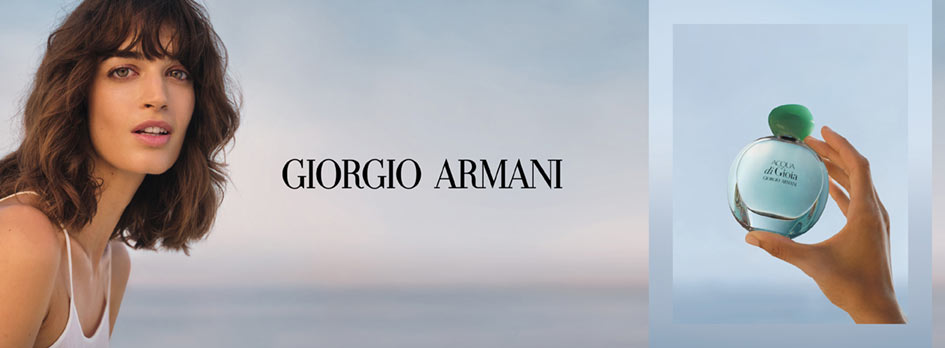 Werbung für Giorgio Armani Acqua di Gioia. Links im Bild ist eine Frau mit schulterlangem braunen Haar, heller Haut und einem weißen Top zu sehen, die vor einem sanften Meeres- oder Himmelhintergrund in die Kamera blickt. In der Mitte steht in schwarzer Schrift 'Giorgio Armani'. Rechts hält eine Hand eine runde, türkisfarbene Parfumflasche mit grünem Verschluss und der Aufschrift 'Acqua di Gioia – Giorgio Armani' in die Höhe.
