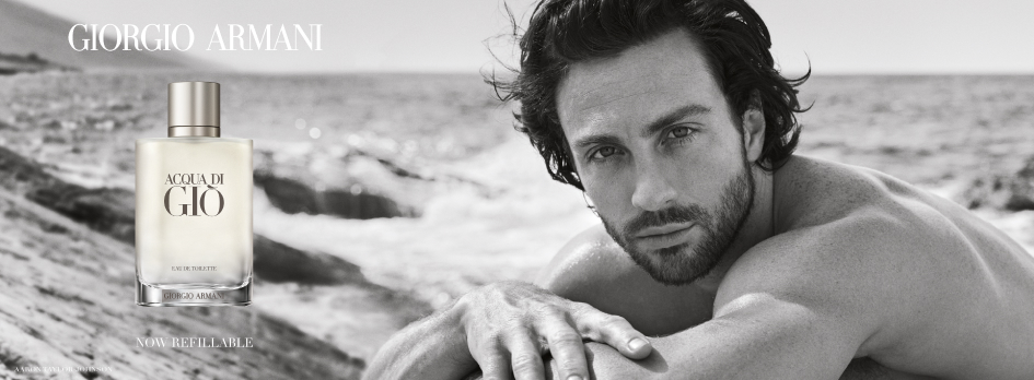 Werbebild für 'Giorgio Armani Acqua di Giò'. In einer schwarz-weißen Strandaufnahme liegt rechts ein durchtrainierter Mann mit dunklem Haar und Bart auf einem Felsen und schaut direkt in die Kamera. Links ist die Parfumflasche mit heller Flüssigkeit und silbernem Verschluss zu sehen. Darüber steht 'Giorgio Armani', darunter 'Now Refillable'.
