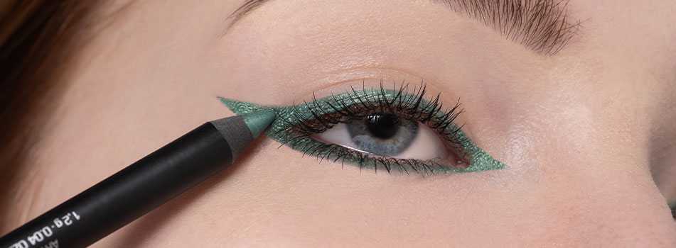 Nahaufnahme eines geschminkten Auges mit einem auffälligen, grünen Eyeliner in Cat-Eye-Form. Ein grüner Kajalstift wird am unteren Wimpernkranz aufgetragen. Die Wimpern sind getuscht, die Iris ist blau.