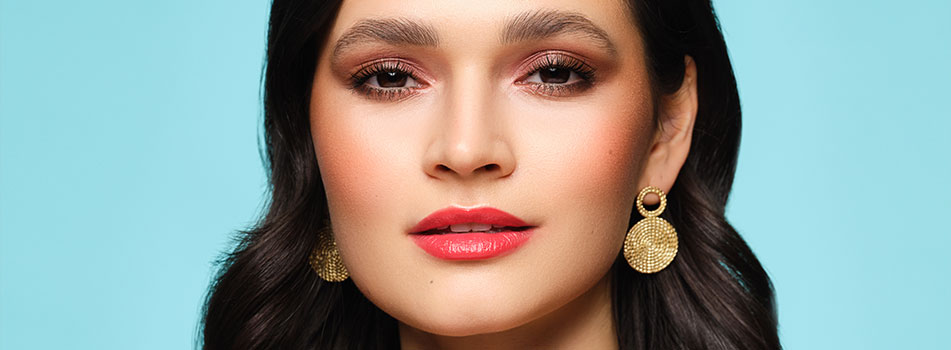 Porträt einer Frau mit elegantem Make-up, rotem Lippenstift und dezentem Lidschatten. Sie trägt auffällige goldene Ohrringe und hat dunkles, gewelltes Haar. Der Hintergrund ist in einem hellen Türkis gehalten.