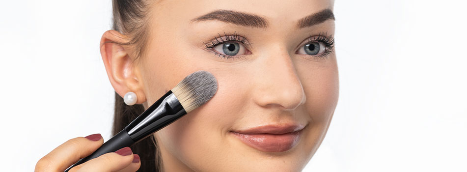 Junge Frau trägt mit einem Foundation-Pinsel Make-up auf ihre Wange auf. Sie hat blaue Augen, braune Haare, Perlenohrringe und ein natürliches Make-up. Der Hintergrund ist weiß und schlicht.