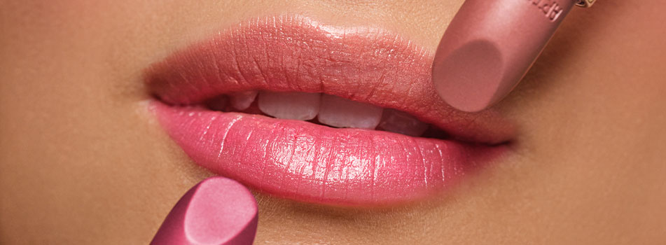Nahaufnahme geschminkter Lippen mit glänzendem pinkfarbenem Lippenstift. Zwei Lippenstifte in Rosatönen werden gleichzeitig an die Lippen geführt. Die Lippen wirken glatt und gepflegt.