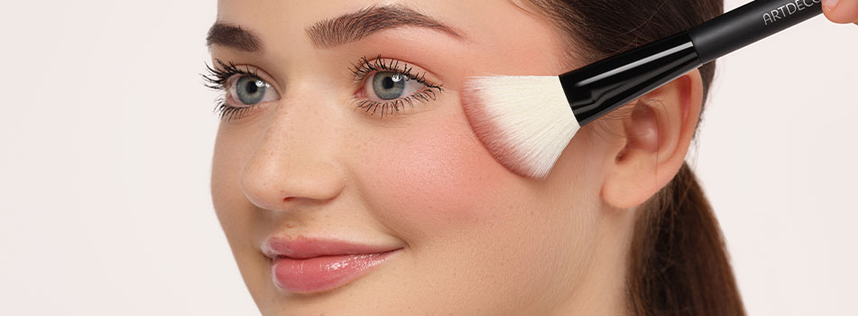 Nahaufnahme einer jungen Frau, die sanft Rouge auf ihre Wange aufträgt. Ein ARTDECO-Pinsel wird zum Auftragen des rosafarbenen Blushs verwendet. Die Frau lächelt dezent und ihre Haut wirkt glatt und gepflegt.