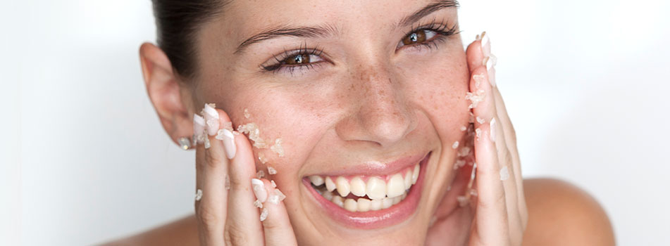 Lächelnde junge Frau mit Peeling-Körnern im Gesicht. Sie reibt sanft ein Gesichtspeeling auf ihre Wangen und schaut direkt in die Kamera. Ihre Haut wirkt frisch und gepflegt.