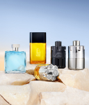 Verschiedene Parfümflakons in unterschiedlichen Farben stehen auf hellem Sand vor einem blauen Himmel. Die Anordnung erinnert an eine elegante Sommerkollektion.