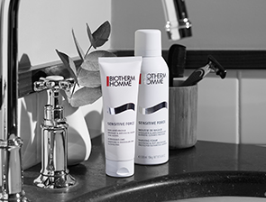Biotherm Homme Rasurpflegeprodukte, darunter Rasierschaum und Aftershave, stehen auf einem Waschbeckenrand neben einem Wasserhahn. Die Szene ist in Schwarz-Weiß gehalten.