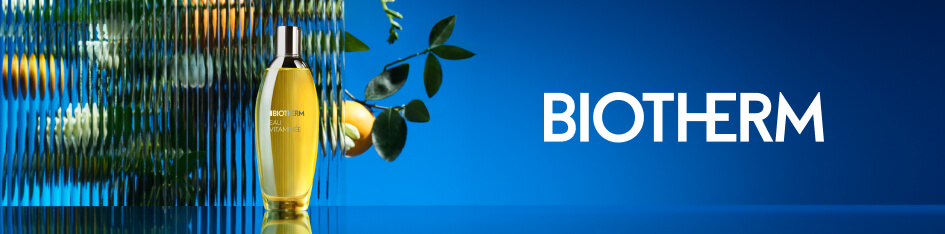 Werbung für Biotherm Eau Vitaminee. Eine gelbe Parfümflasche steht auf spiegelndem Untergrund vor einer geriffelten Glasfläche mit Zitrusfrüchten und Blättern im Hintergrund. Rechts befindet sich der Markenname 'Biotherm' auf blauem Farbverlauf.