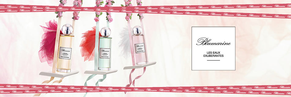 Drei Parfumflakons der Blumarine-Kollektion „Les Eaux Exubérantes“ in Rosa-, Grün- und Pfirsichtönen hängen an rosa Schleifen mit Tüllverzierung. Im Hintergrund steht das Blumarine-Logo mit der Aufschrift „Les Eaux Exubérantes“.