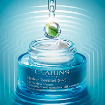 Clarins Hydra-Essentiel [HA²] Tagescreme in einem blauen Glastiegel mit weißem Deckel. Darüber schwebt ein Tropfen mit grün-blauer Farbwirbelung, eingefangen in einer Pipette. Der Hintergrund ist in leuchtendem Blau mit kreisförmigen Lichtwellen gestaltet.