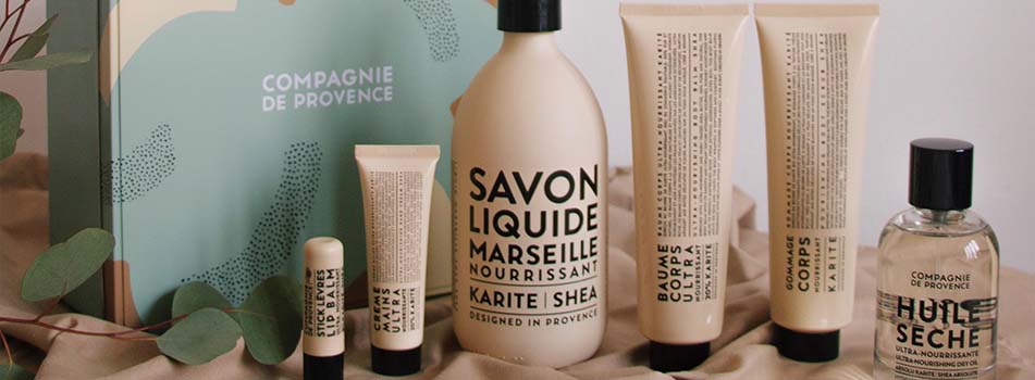 Pflegeserie von Compagnie de Provence mit Karité Shea: verschiedene Produkte wie Flüssigseife, Handcreme, Lippenbalsam, Körperbalsam, Körperpeeling und ein Trockenöl stehen nebeneinander vor einer Geschenkbox mit Pflanzenmotiven.
