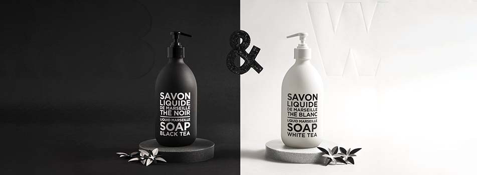 Zwei Flüssigseifenflaschen von Compagnie de Provence stehen nebeneinander – links eine schwarze Flasche mit schwarzem Pumpspender und der Aufschrift 'SOAP BLACK TEA', rechts eine weiße Flasche mit weißem Pumpspender und der Aufschrift 'SOAP WHITE TEA'. Die Flaschen stehen jeweils auf runden Podesten vor schwarzem bzw. weißem Hintergrund. Dazwischen befindet sich ein schwarzes '&'-Symbol.