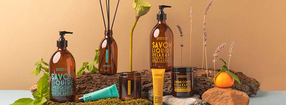 Pflegeprodukte von Compagnie de Provence in bernsteinfarbenen Verpackungen arrangiert auf Baumrinde mit Naturdekoration. Zu sehen sind unter anderem Flüssigseifen mit den Aufschriften 'Energisant' und 'Relaxant', Körpercreme, Handcreme sowie ein Duftstäbchen-Glasflakon. Im Hintergrund Lavendel, ein Mandarinchen, Moose und Blumen.
