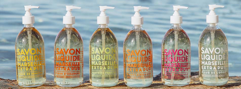 Sechs transparente Flaschen Flüssigseife von Compagnie de Provence stehen nebeneinander auf einer Natursteinfläche am Wasser. Jede Flasche trägt farbige Aufschriften wie 'Fleur de Vigne', 'Fleur d'Oranger', 'Verveine Fraîche', 'Pamplemousse', 'Bois Sauvage' und 'Fleur de Coton'.