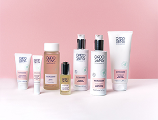 Eine Auswahl an Hautpflegeprodukten auf einem hellrosa Hintergrund. Die Serie ist speziell für trockene Haut und Neurodermitis konzipiert. Zu sehen sind Cremes, Fluids und ein Serum in weißen und rosafarbenen Verpackungen.