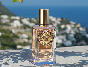 Parfumflakon von Dolce & Gabbana Devotion mit goldener Kappe und herzförmigem Emblem steht auf einem hellen Steinpodest. Im Hintergrund ein mediterranes Küstenpanorama mit blauem Meer, weißen Häusern und grüner Landschaft.