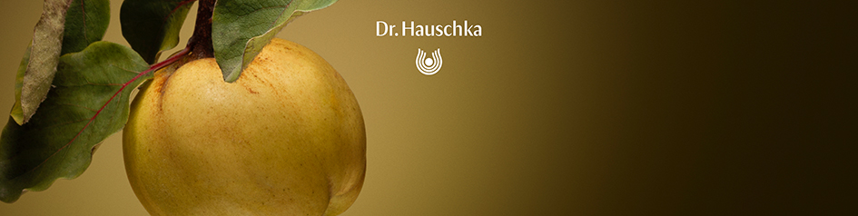 Werbebild von Dr. Hauschka: Nahaufnahme einer goldgelben Quitte mit grünen Blättern vor einem warmen braun-goldenen Hintergrund. Darüber steht das Dr. Hauschka-Logo in Weiß – das Motiv symbolisiert natürliche Pflege und Frische.