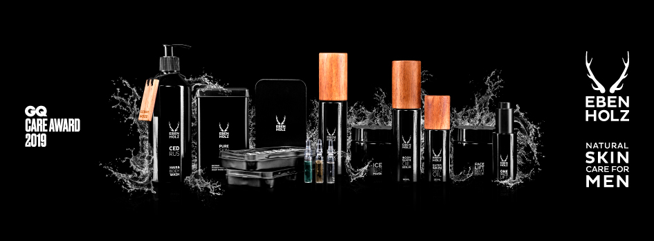 Werbebild für EBENHOLZ Natural Skin Care for Men: Eine Reihe schwarzer Pflegeprodukte mit Holzakzenten steht vor schwarzem Hintergrund, umgeben von spritzendem Wasser. Links das „GQ Care Award 2019“-Logo, rechts das Markenlogo mit Hirschgeweihsymbol und dem Schriftzug „EBENHOLZ – Natural Skin Care for Men“.