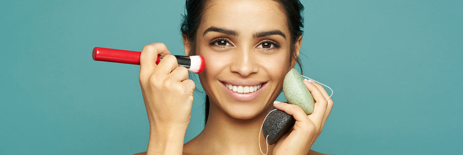 Lächelnde Frau mit glatter Haut vor türkisfarbenem Hintergrund, trägt mit einem roten Pinsel Make-up auf und hält zwei Konjac-Schwämme in der anderen Hand.