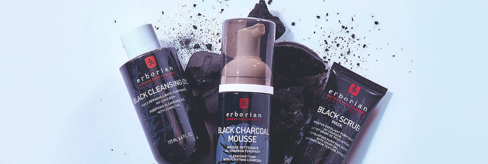Drei Reinigungsprodukte von Erborian mit Aktivkohle: Black Cleansing Oil, Black Charcoal Mousse und Black Scrub Mask, arrangiert mit Kohlestücken auf hellem Hintergrund.