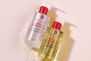 Zwei Pumpflaschen Erborian Centella Cleansing Gel und Cleansing Oil auf pastellfarbenem Hintergrund, mit sichtbarem Schaum um die Produkte.