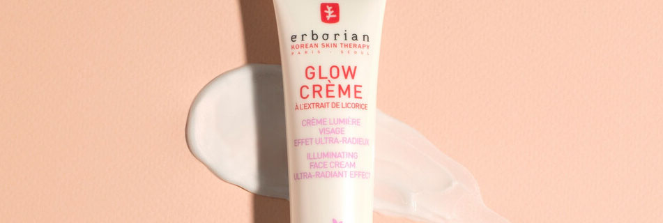 Erborian Glow Crème in einer weißen Tube mit roter Schrift, stehend auf pfirsichfarbenem Hintergrund mit aufgetragener, schimmernder Produkttextur.