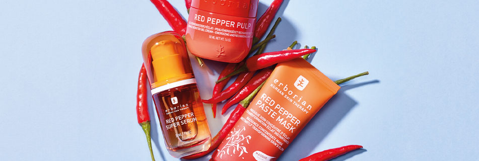 Drei Gesichtspflegeprodukte aus der Red Pepper-Serie von Erborian – Super Serum, Pulp und Paste Mask – dekorativ arrangiert mit frischen roten Chilis auf hellblauem Hintergrund.
