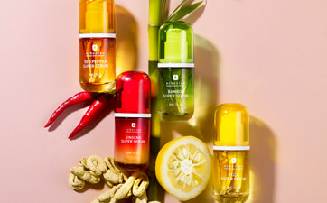 Vier bunte Fläschchen der Marke Erborian mit den Varianten Red Pepper, Bamboo, Ginseng und Yuzu Super Serum, arrangiert mit passenden natürlichen Zutaten wie Chilis, Bambus, Ginseng und Zitrone auf pastellfarbenem Hintergrund.