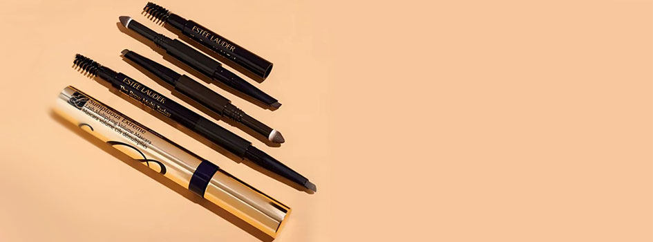 Verschiedene Estée Lauder Augen-Make-up-Produkte, darunter zwei Augenbrauenstifte mit Bürstchen, ein Lidschattenstift und eine goldene Sumptuous Extreme Mascara, arrangiert auf einem pfirsichfarbenen Hintergrund.