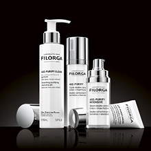 Vier Filorga Pflegeprodukte aus der AGE-PURIFY Serie in weiß-silberner Verpackung, auf einem glänzenden schwarzen Untergrund stehend. Enthalten sind ein Reinigungsgel mit Pumpspender, zwei Seren und eine Creme.
