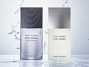 Zwei Parfumflakons aus der Serie 'L’Eau d’Issey Pour Homme' von Issey Miyake stehen nebeneinander auf einer spiegelnden Wasserfläche. Links die Variante 'Solar Lavender' in einem transparent-lila Flakon mit dunklem Holz-Look-Verschluss, rechts die klassische Version in einem milchig-weißen Flakon mit silbernem Verschluss. Im Hintergrund spritzt Wasser, was Frische und Klarheit vermittelt.