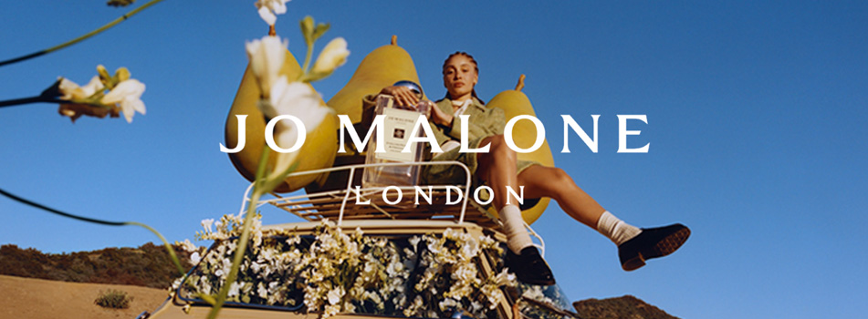 Werbebild von Jo Malone London: Eine Person sitzt auf einem mit weißen Blüten und übergroßen Birnen geschmückten Fahrzeug vor blauem Himmel und Landschaft im Hintergrund. In der Mitte der Schriftzug „Jo Malone London“.