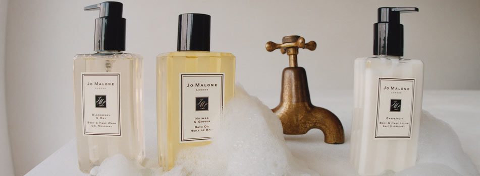 Drei Pflegeprodukte von Jo Malone London stehen neben einem alten Messingwasserhahn inmitten von Schaumblasen. Die Flaschen enthalten Body & Hand Wash, Bath Oil und Body & Hand Lotion.