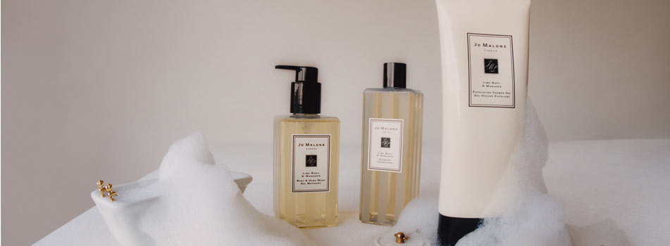Drei Pflegeprodukte von Jo Malone London stehen auf einer hellen Fläche zwischen Seifenschaum und goldenen Accessoires. Zu sehen sind Body & Hand Wash, Bath Oil und eine große Tube Body Crème.