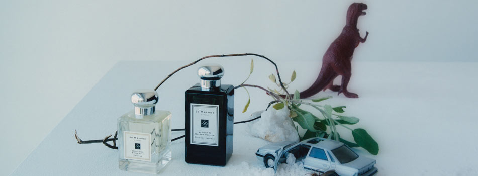 Zwei Parfumflakons von Jo Malone London stehen auf einer hellen Fläche neben einem kleinen Ast. Dahinter befinden sich ein Spielzeugauto, etwas Grünpflanze und eine kleine Dinosaurierfigur.