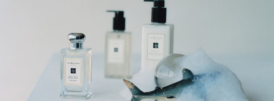 Jo Malone London Produkte, darunter ein Parfumflakon und eine Lotion mit Pumpspender, stehen auf heller Fläche neben einem Seifenblasen-Schaum und einer kleinen Muschel.
