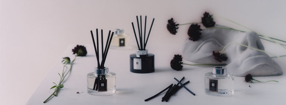 Mehrere Duftdiffusoren von Jo Malone London mit schwarzen Stäbchen stehen auf einer weißen Fläche, umgeben von dunklen Blüten und einer weißen, skulpturalen Gesichtsform.