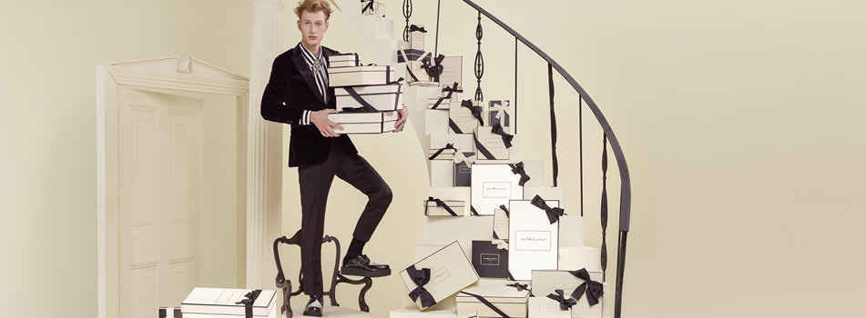 Ein Mann in elegantem Anzug steht auf einer Treppe voller cremefarbener Geschenkboxen mit schwarzen Schleifen von Jo Malone London und hält mehrere davon in den Armen.