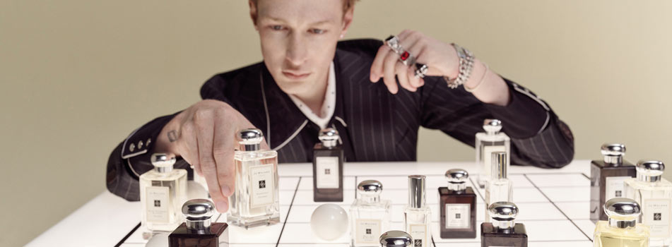 Eine Person in dunklem Anzug arrangiert mehrere Parfumflakons von Jo Malone London auf einer weiß gekachelten Fläche, vor neutralem Hintergrund.