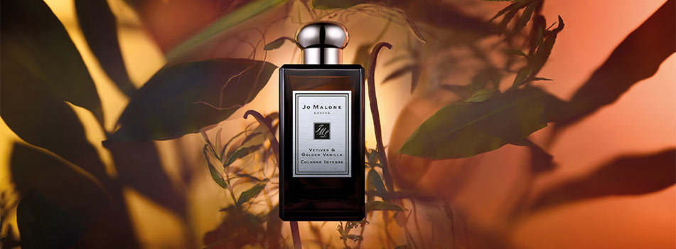 Dunkler Flakon des Dufts „Vetiver & Golden Vanilla Cologne Intense“ von Jo Malone London vor warmem orange-braunem Hintergrund mit Blättern und sanftem Lichtspiel.
