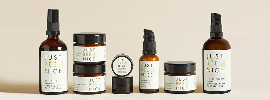 Mehrere Pflegeprodukte der Marke Just Bee Nice stehen nebeneinander auf einer hellen Fläche. Die braunen Glasflaschen und Tiegel tragen weiße Etiketten mit schwarzer und goldener Schrift.