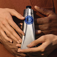 Zwei Hände halten gemeinsam eine silberne Tube L'OCCITANE Handcreme.